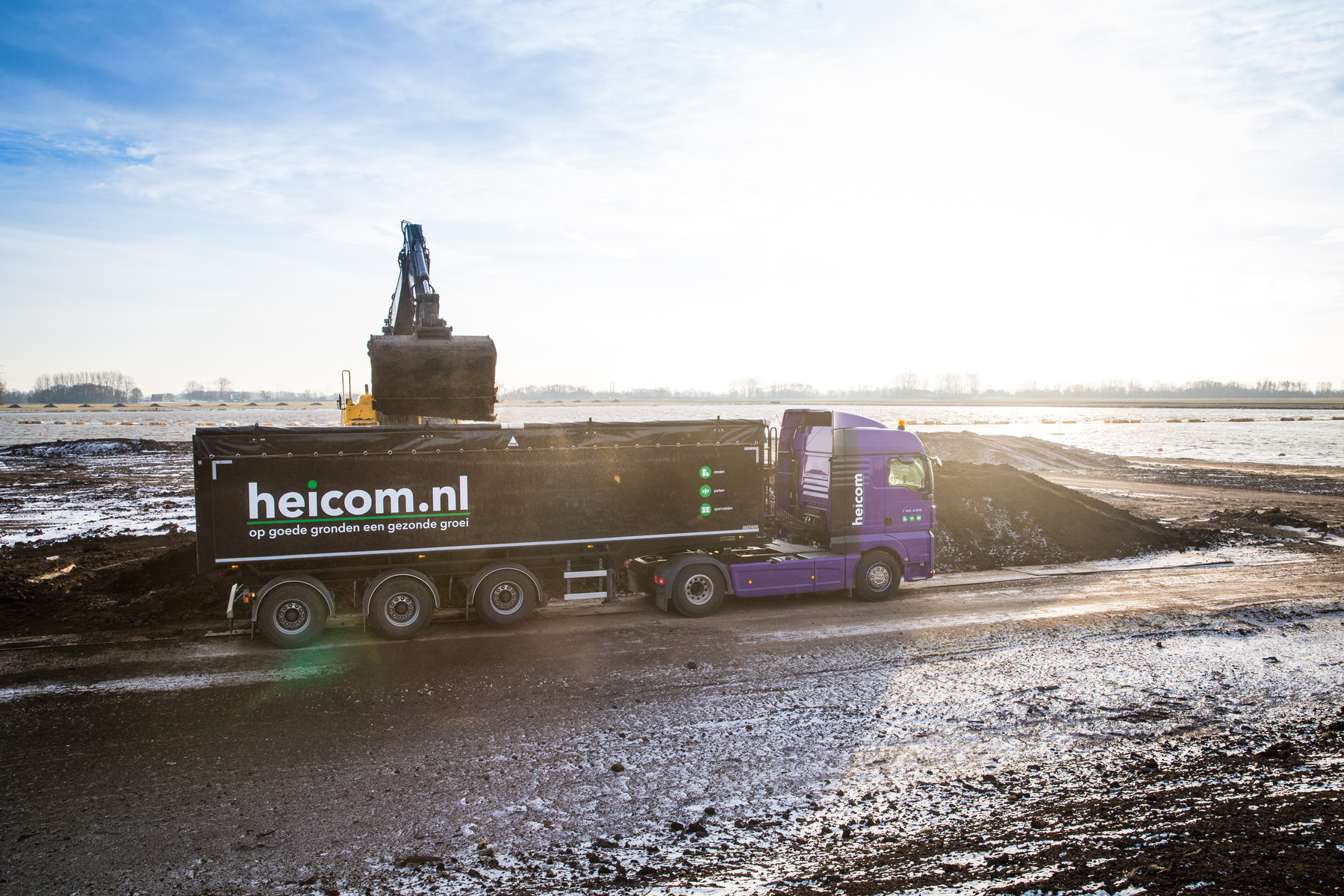 Graafmachine laadt een oplegger van Heicom op een nat werkterrein