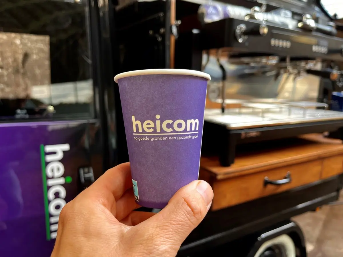 Hand houdt een paarse heicom-koffiebeker vast bij een espressobar