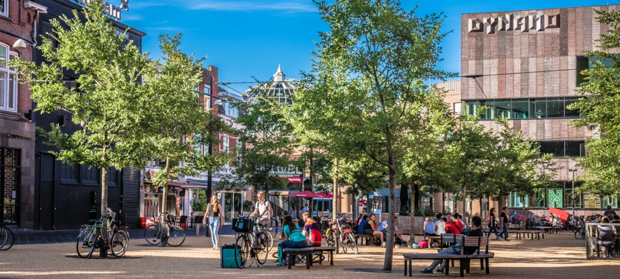 Plein met jonge bomen, bankjes en fietsers voor het gebouw Dynamo