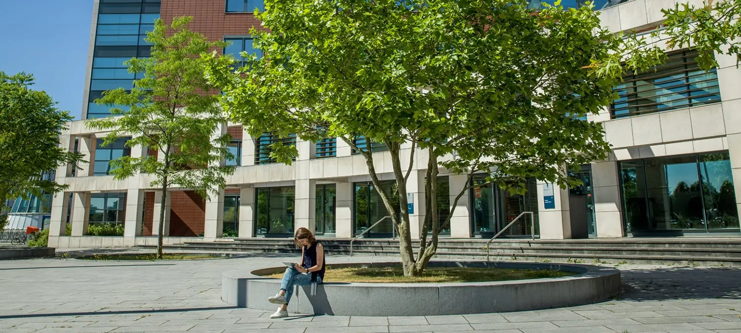 Vrouw zit op een betonnen bank onder een boom bij een kantoorgebouw