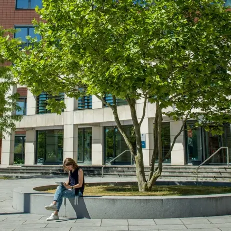 Vrouw zit op een betonnen bank onder een boom bij een kantoorgebouw