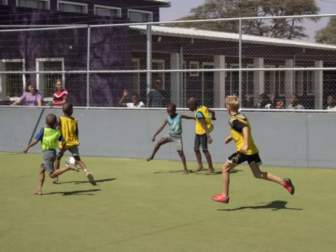 Kinderen in hesjes voetballen op een kunstgrasveld achter een hek