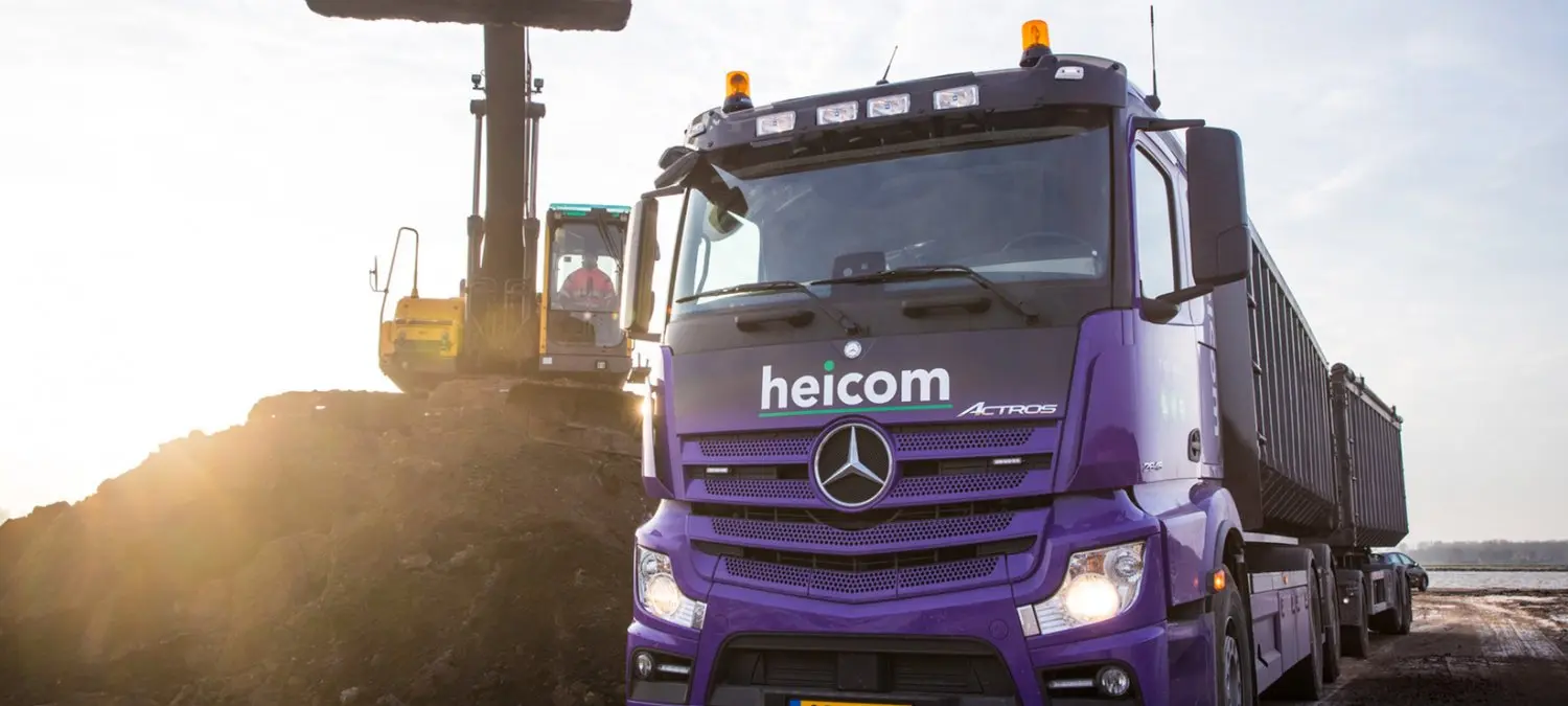 Paarse Heicom-vrachtwagen bij een gronddepot met graafmachine in tegenlicht
