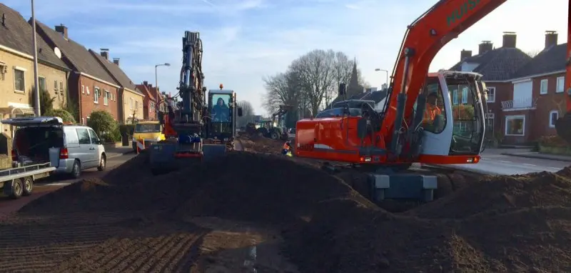 Twee oranje graafmachines werken in een opengebroken straat met grondhopen