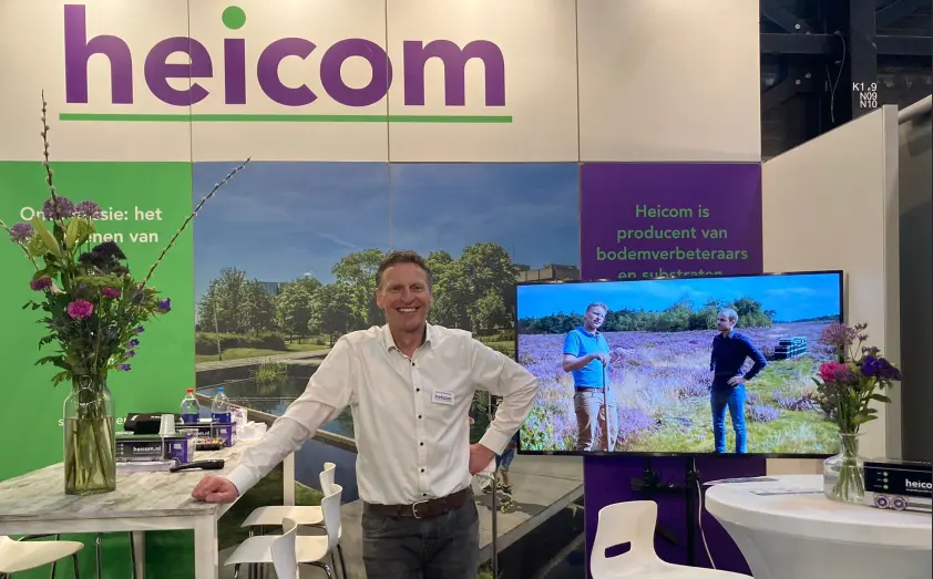 Lachende man bij de beursstand van Heicom met bloemen en beeldscherm
