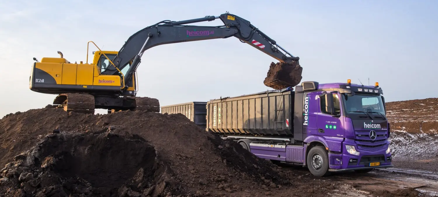 Graafmachine laadt donkere grond in de container van een paarse Heicom-vrachtwagen