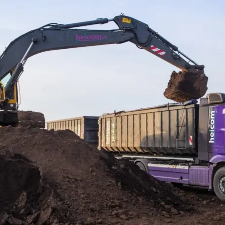 Graafmachine laadt donkere grond in de container van een paarse Heicom-vrachtwagen