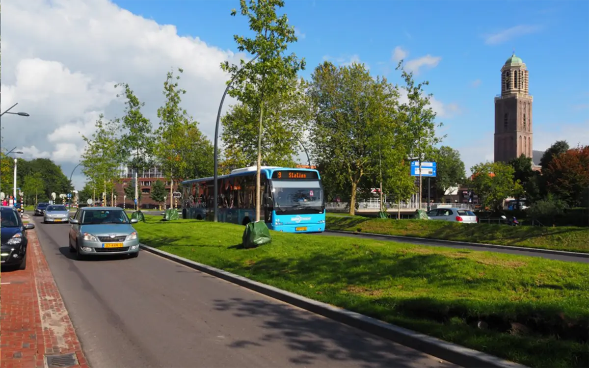Stadsweg met grasberm en jonge bomen, blauwe stadsbus rijdt langs een kerktoren