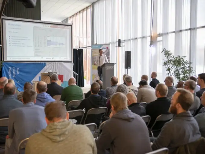 Zaal met toehoorders kijkt naar een spreker bij een presentatiescherm