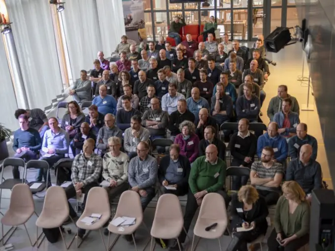 Volle zaal met deelnemers op stoelen, gezien van bovenaf