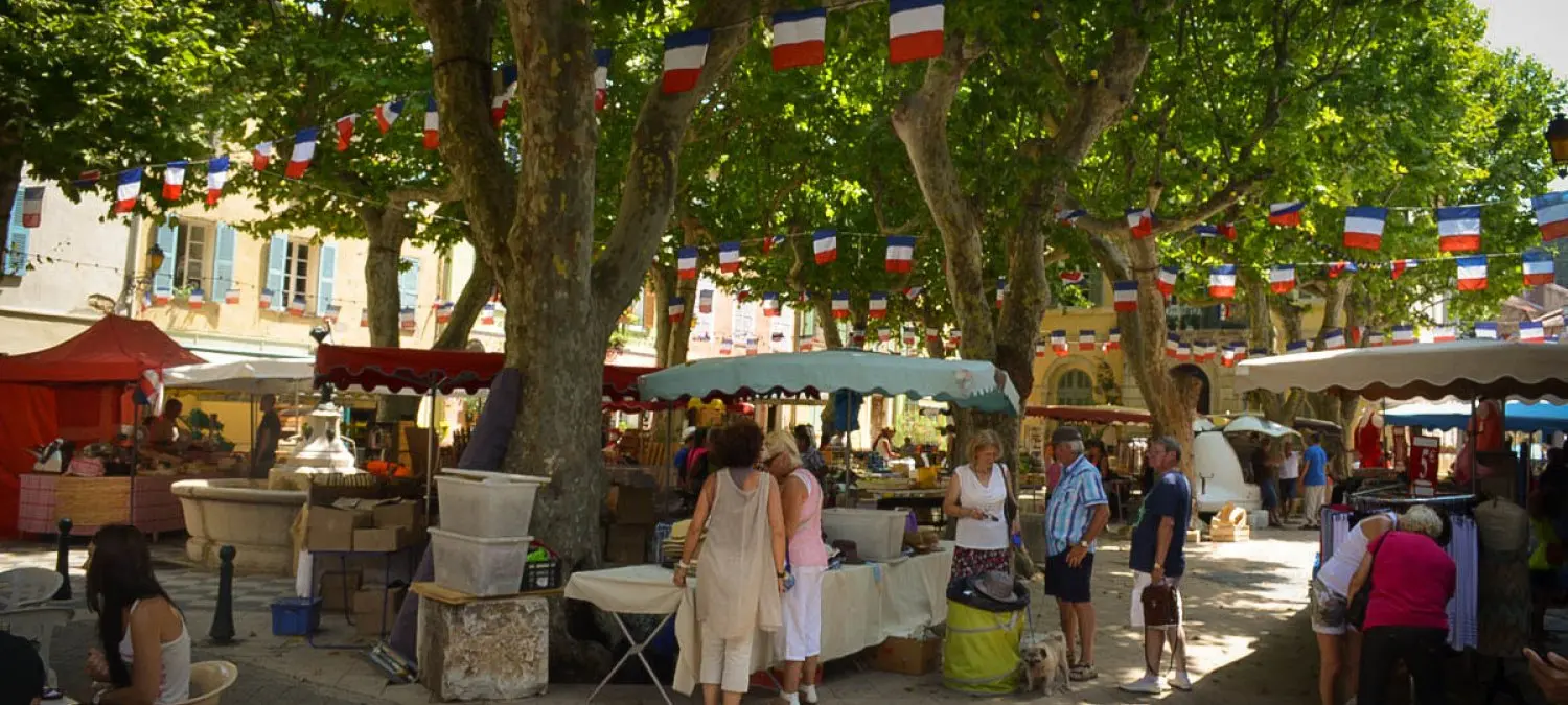 Marktkraampjes onder platanen op een Frans dorpsplein met vlaggetjes