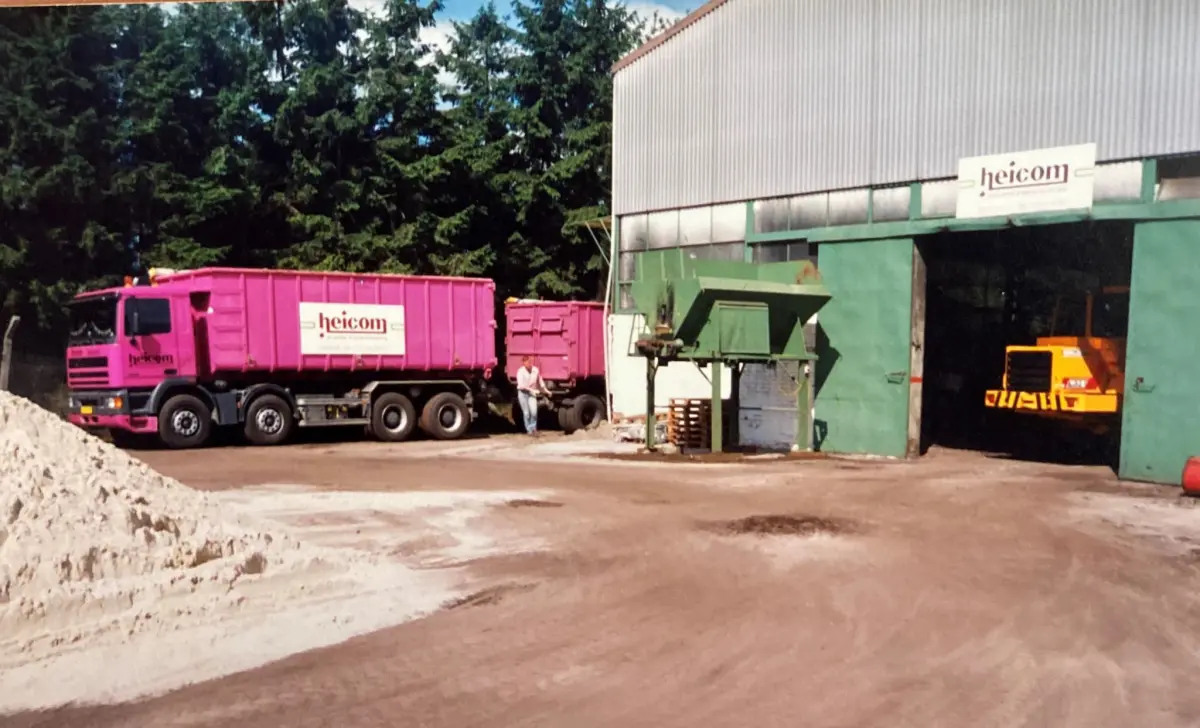Roze Heicom-truck met aanhanger bij een loods en een hoop zand
