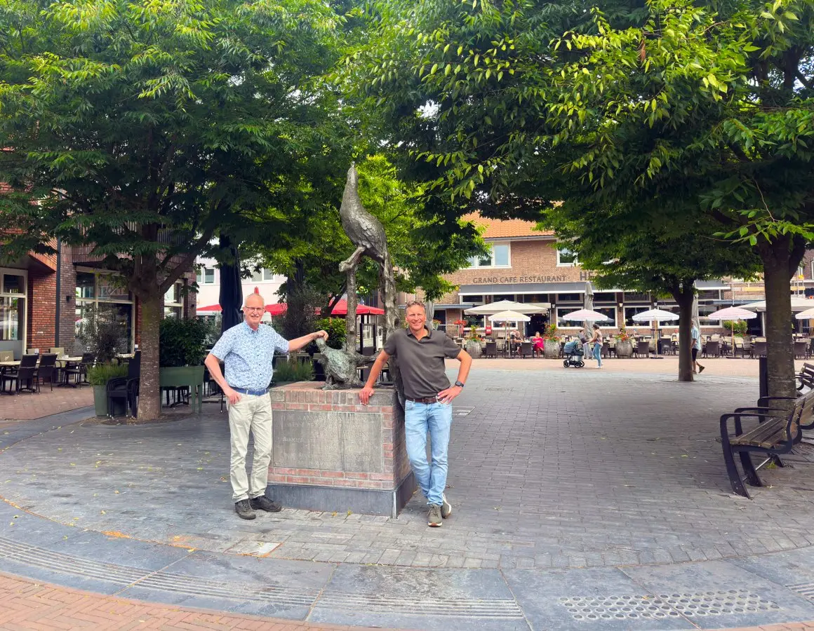 Twee mannen bij een standbeeld op een bestraat plein met bomen