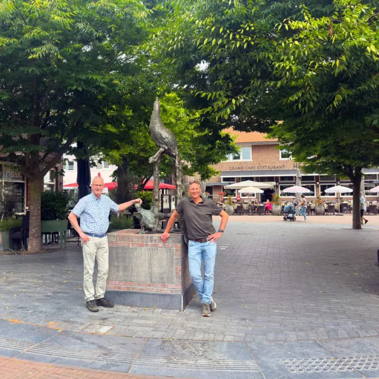 Twee mannen bij een standbeeld op een bestraat plein met bomen