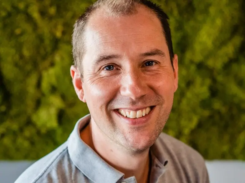 Portret van Joeri Faas in grijze polo, lachend voor een mosmuur