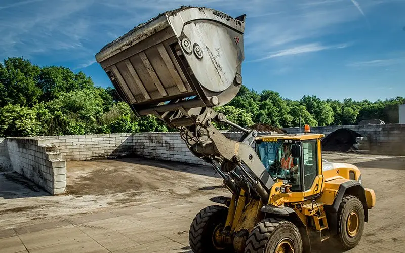 Shovel met geheven bak op een buitenterrein langs opslagvakken