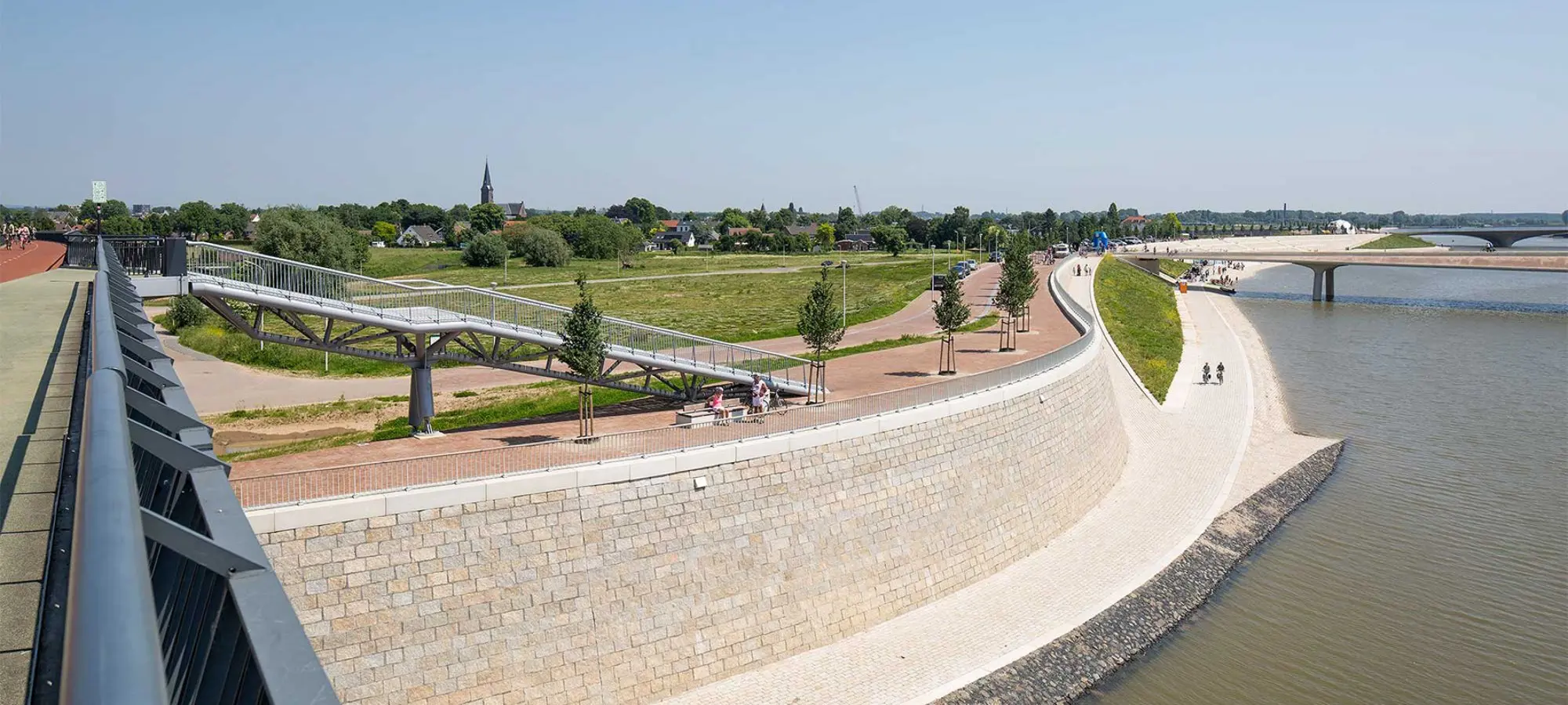 Wandelpromenade langs een rivier met bomenrij en brug in de verte