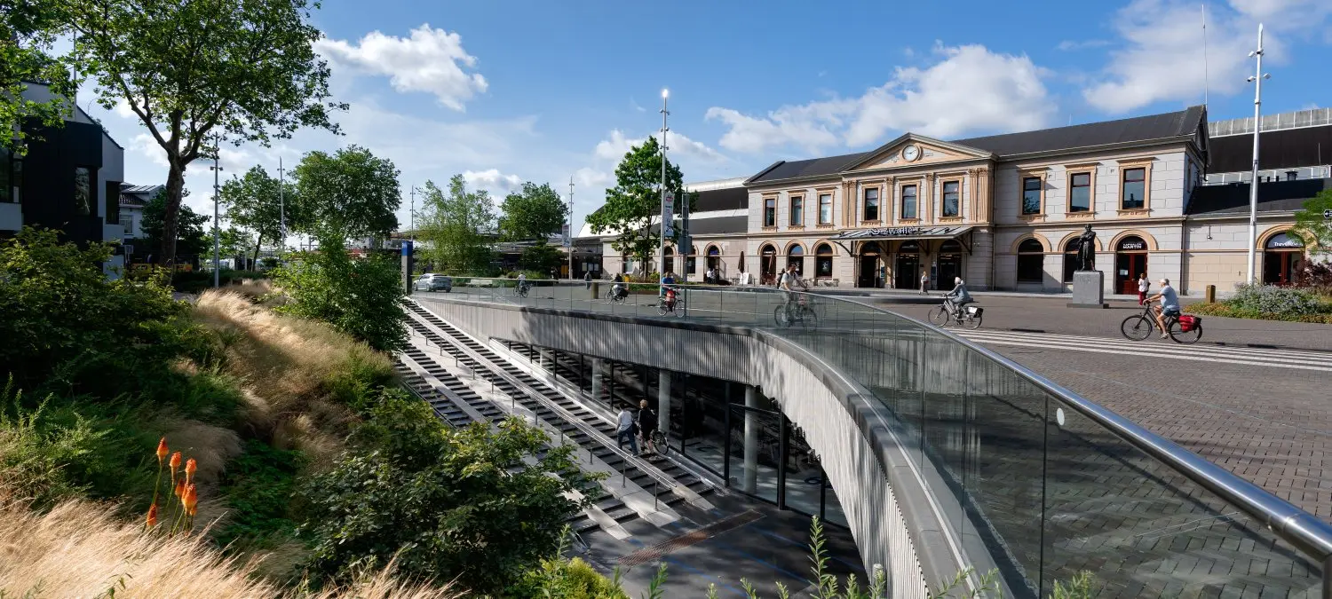 Stationsplein met fietsers en een verdiepte fietsenstalling voor een oud stationsgebouw