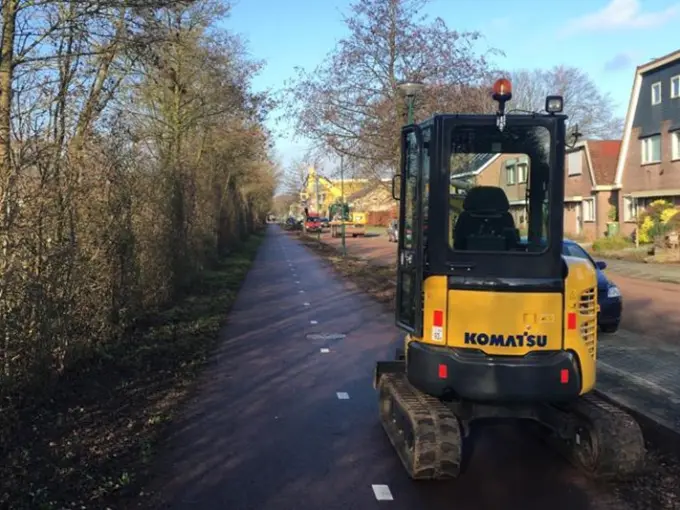 Minigraafmachine op een fietspad tussen kale bomen in een woonstraat