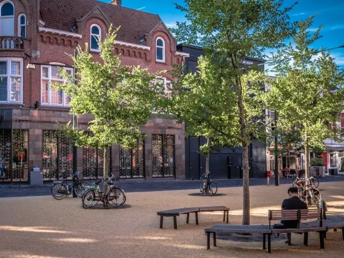 Man op een bank onder jonge bomen bij een bakstenen hoekpand