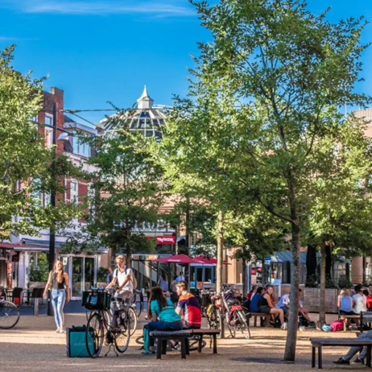 Plein met jonge bomen, bankjes en fietsers voor het gebouw Dynamo
