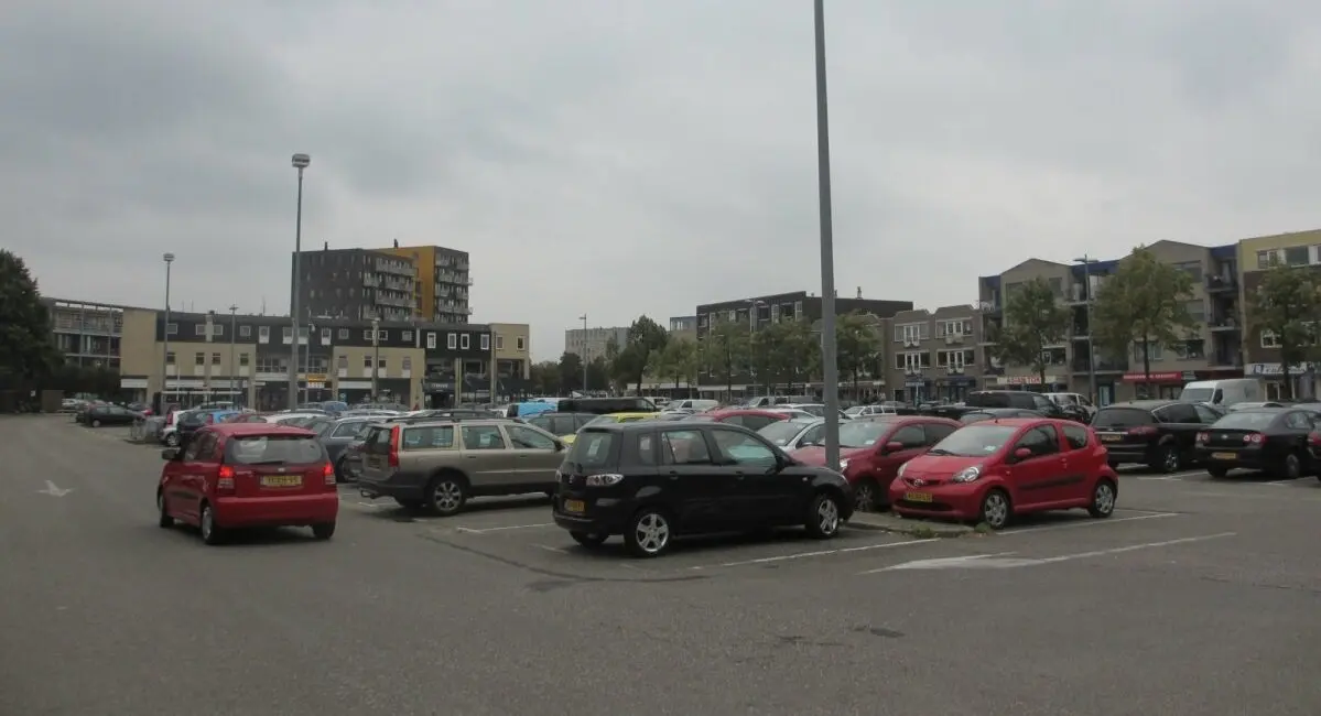 Volle parkeerplaats met auto's voor winkels en woningen onder een grijze lucht