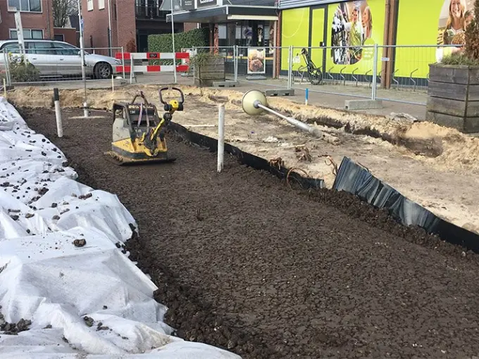 Trilplaat verdicht donkere grond in een sleuf langs een winkelpui