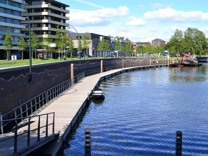 Houten vlonderpad langs een kademuur met appartementen en jonge bomen