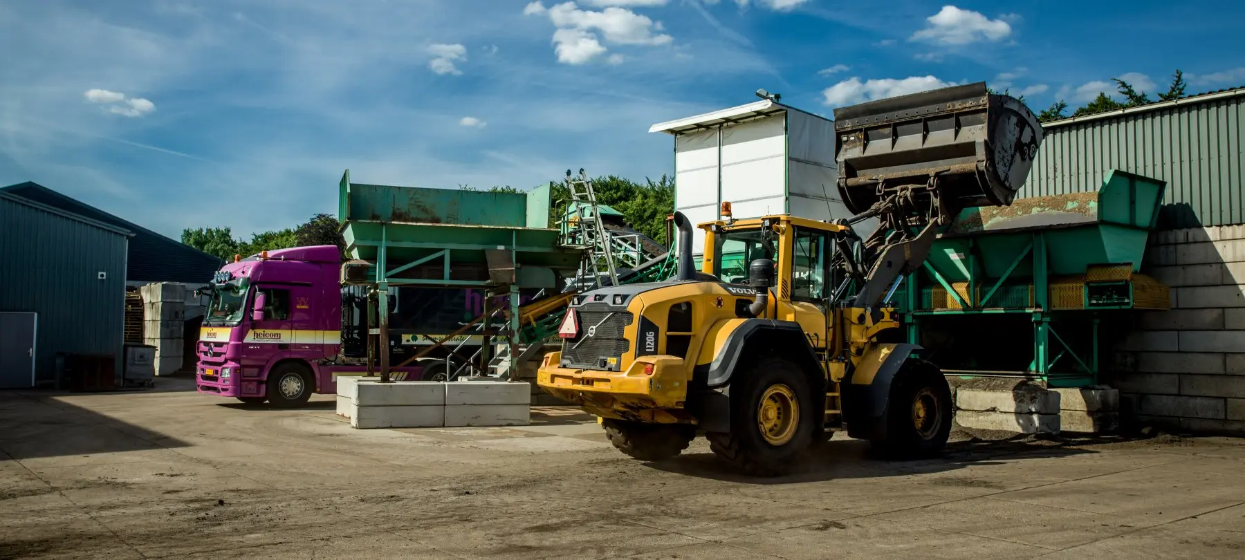 Shovel met geheven bak naast een vrachtwagen en zeefinstallatie op een bedrijfsterrein