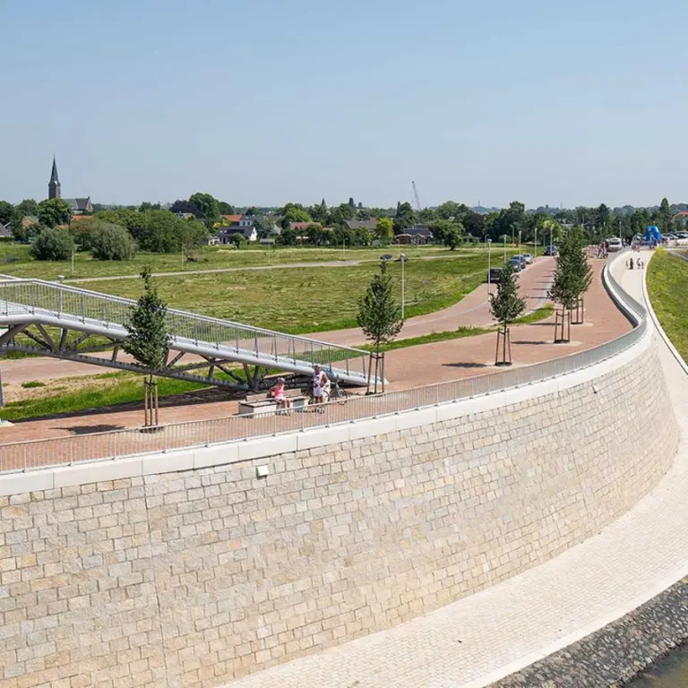 Wandelpromenade langs een rivier met bomenrij en brug in de verte