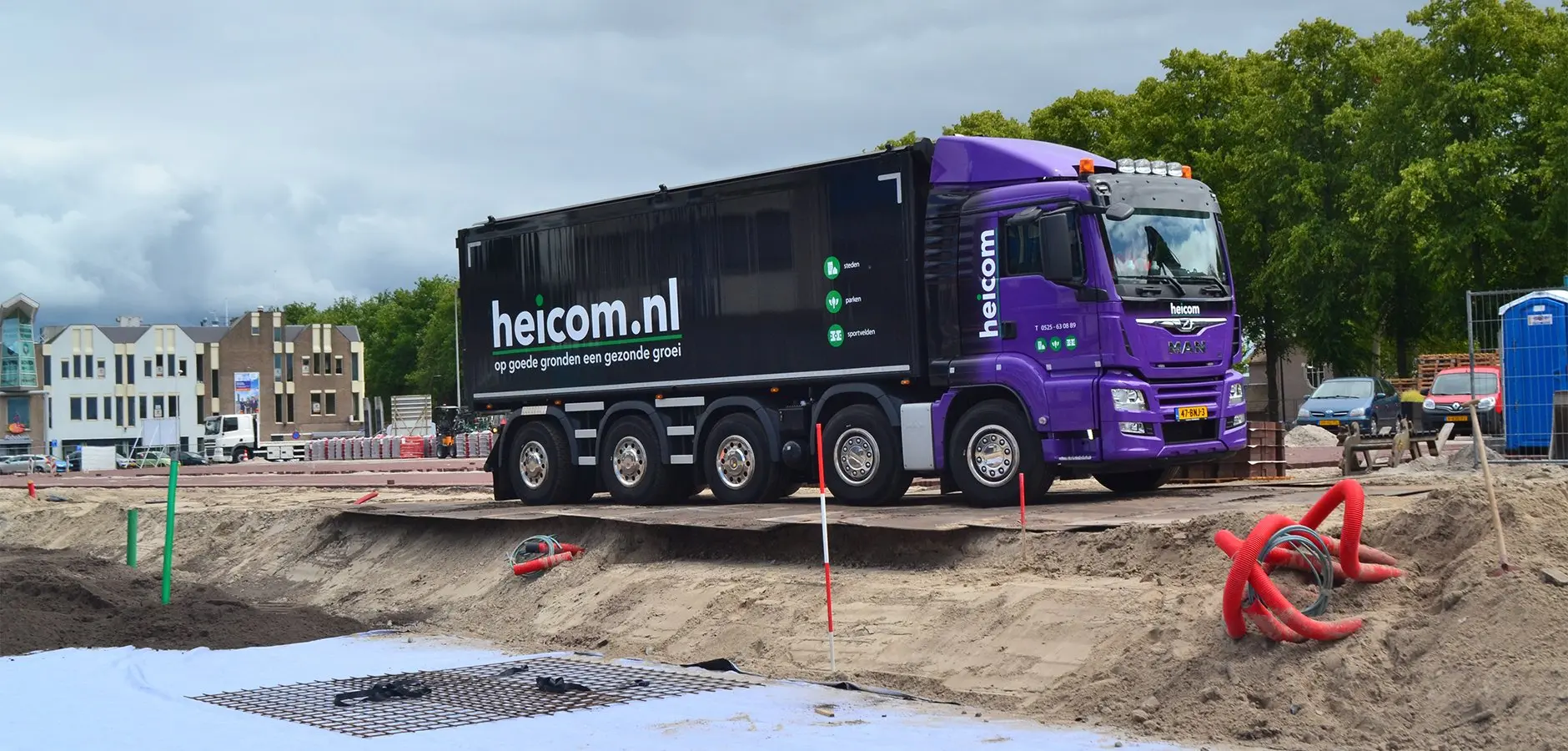 Paarse vrachtwagen met het opschrift heicom.nl op een bouwterrein