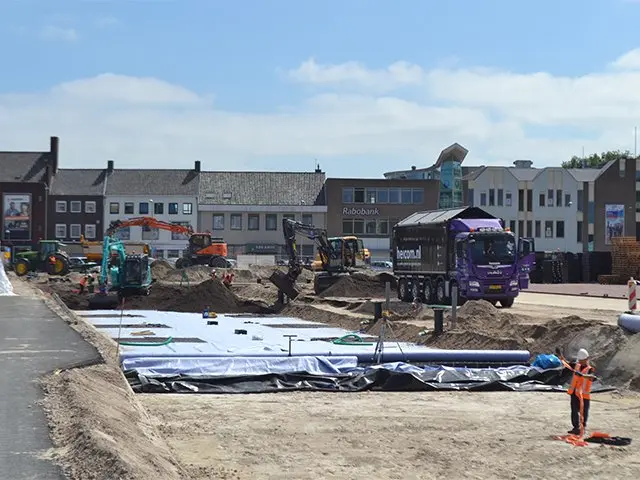 Graafmachines en een Heicom-vrachtwagen op een bouwplaats aan een dorpsplein