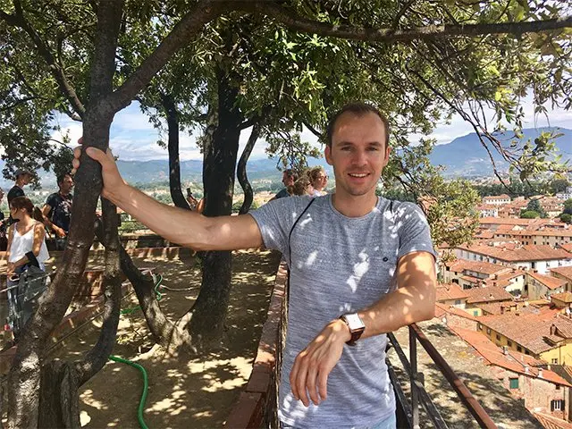 Man leunt op een reling onder bomen met uitzicht over rode daken